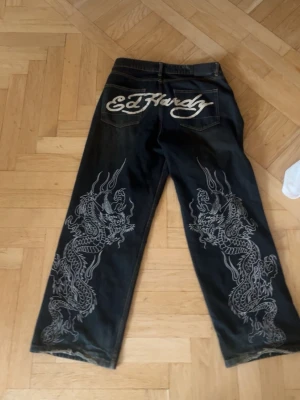 Ed Hardy jeans med drakmotiv - Säljer ett par svarta Ed Hardy jeans med bred passform och stora vita drakmotiv längs båda benen. Bakfickan har en stor Ed Hardy-logga i vitt och framtill finns en mindre logga. 