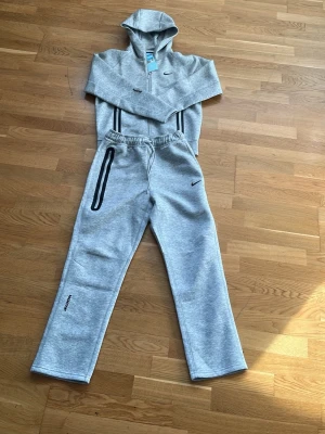 Grå mjukisbyxor från Nike NOCTA - Snygga grå mjukisbyxor från Nike NOCTA med svarta detaljer och logga på benet. Byxorna har resår i midjan och raka ben, samt en markerad ficka med dragkedja. Perfekta för en avslappnad och sportig stil.