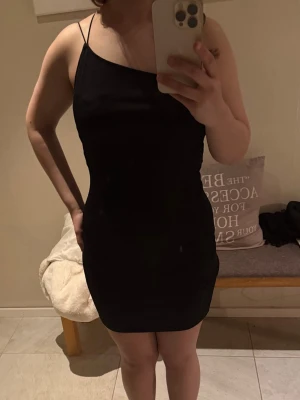 Svart kort klänning från H&M - Säljer en svart, tight kortklänning med tunna axelband och rak halsringning. Klänningen har en enkel och stilren design som sitter figurnära och är perfekt för dig som gillar minimalistisk stil. Materialet känns mjukt och stretchigt.