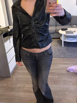 Svart croppad skjorta i skinnimitation - Säljer en svart croppad skjorta i skinnimitation med lång ärm och knappar framtill. Skjortan har en snygg rynkad detalj längs knappraden och en djup v-ringning. Perfekt för dig som vill ha en edgy och trendig look.