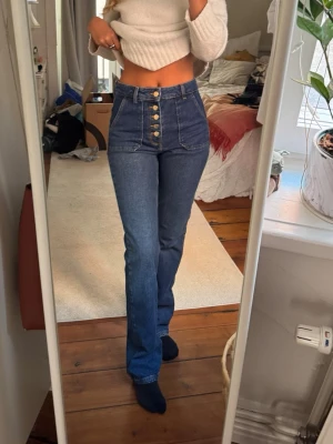 Blå high waist bootcut jeans med knappar - Säljer ett par blå high waist jeans med utsvängda ben och fem synliga guldknappar framtill. Jeansen har stora fickor fram och är i klassisk denim. Perfekta för en trendig och avslappnad look.❤️⭐️