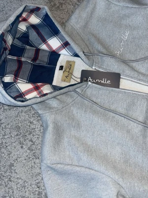 Grå hoodie från Aumille  - Snygg grå hoodie från Aumille med dragkedja och fickor framtill. Huvan har ett rutigt foder i blått, vitt och rött som ger en cool kontrast. Broderad logga på bröstet och ribbade muddar vid ärmar och midja. Perfekt för en avslappnad och trendig look. Köpt för 1400kr men går för 850. Helt oanvänd,  lappen finns kvar!