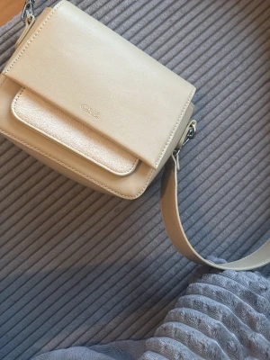 Beige axelremsväska från CW - Prislappen är fortfarande på nypriset är 400krStilren beige handväska från CW med axelrem och lock. Väskan har en fyrkantig form och en mindre ficka framtill med metallic-detalj. Tillverkad i slätt syntetmaterial och har diskret logga på framsidan. Pris går att diskuteras vill bli av med den