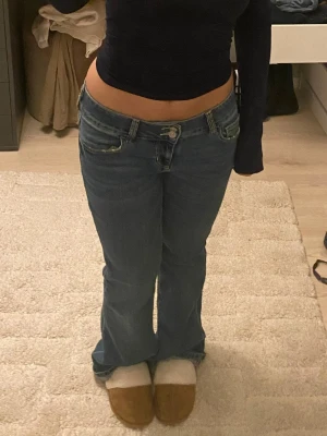 Blå lowwaist bootcut jeans  - Säljer ett par blå bootcut jeans med snygg broderad detalj på bakfickan och knappstängning. Jeansen har låg midja och klassisk femficksdesign. Perfekta för dig som gillar en avslappnad men trendig look.