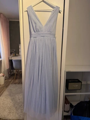Ljusblå balklänning med omlott - Säljer en ljusblå långklänning med omlottdesign och djup v-ringning. Klänningen har breda axelband och ett markerat midjeband som knyts i en stor rosett bak. Tillverkad i ett tunt, flygigt tyllmaterial som ger en romantisk känsla.