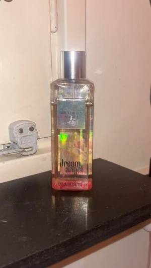 Victoria's Secret Dream Angel - Säljer en Victoria's Secret Dream Angel parfym i en genomskinlig, fyrkantig flaska med holografiska detaljer och silverfärgat lock. Flaskan rymmer 250 ml och har en snygg regnbågsskimrande etikett som ger ett lyxigt intryck. Använd, ungefär 80% kvar.