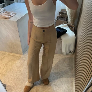 Beige kostymbyxa med hög midja - Snygga beige kostymbyxor med hög midja och raka ben. Byxorna har två framfickor, bälteshällor och stängs med knapp och dragkedja. Perfekta för en stilren och trendig look.