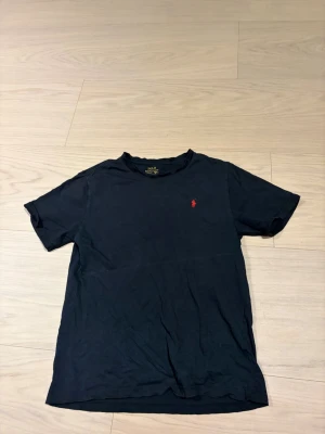Svart t-shirt från Polo Ralph Lauren - Klassisk svart t-shirt från Polo Ralph Lauren med röd broderad logga på bröstet. T-shirten har rund hals och korta ärmar, tillverkad i mjuk bomull för en skön känsla.