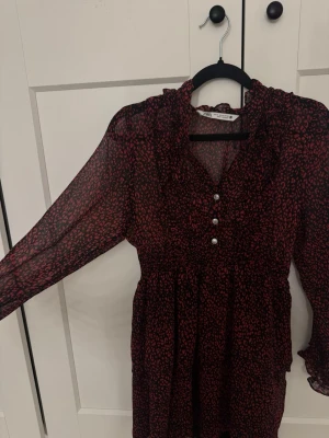 Svart och röd klänning från Zara - Säljer en svart och röd klänning från Zara med blommigt mönster och pärlknappar framtill. Klänningen har långärmade transparenta ärmar, volangdetaljer vid halsen och en v-ringad front. Perfekt för dig som gillar trendiga detaljer.
