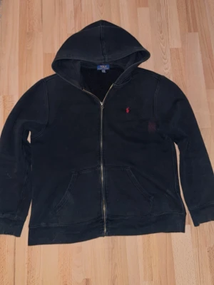 Svart hoodie från Polo Ralph Lauren - Klassisk svart hoodie från Polo Ralph Lauren med dragkedja framtill och huva. Tröjan har kängurufickor och den ikoniska röda loggan broderad på bröstet. Tillverkad i mjuk bomullsblandning som är skön att bära. Perfekt för en avslappnad och stilren look.