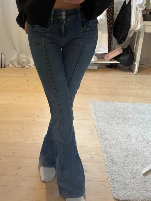 Blå bootcut jeans med fickor fram - Säljer ett par blå bootcut jeans med markerade sömmar och stora fickor framtill. Jeansen har hög midja och stängs med knapp och dragkedja. Perfekta för en trendig och avslappnad look. Materialet är klassisk denim i bomull.
