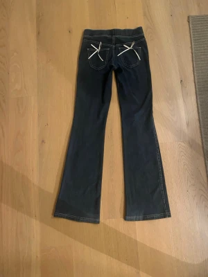 Bootcut jeans med rosetter från H&M - Mörkblå bootcut jeans från H&M i storlek 152. Byxorna har vita kontrastsömmar och söta rosetter på bakfickorna. Tillverkade i mjukt bomullsmaterial med fejk fickor på framsidan och bekväm midja.