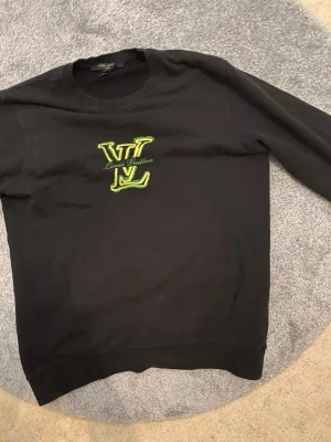Svart Louis Vuitton sweatshirt - Cool svart sweatshirt från Louis Vuitton med stort LV-logo broderat i grönt och gult på bröstet. Tröjan har rund halsringning och långa ärmar. Perfekt för dig som gillar streetwear och vill sticka ut med en exklusiv look.