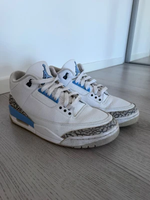 Air Jordan 3 Retro UNC sneakers - Säljer ett par Air Jordan 3 Retro UNC sneakers i vitt läder med ljusblå och grå detaljer. Skorna har den klassiska elefantmönstrade panelen, Jumpman-logga på plösen och AIR-text på hälen. Snygg siluett och ikonisk design som sticker ut.