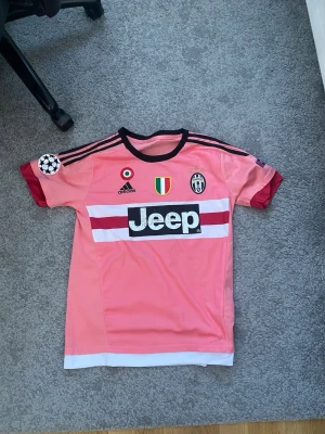 Juventus rosa Pogba tröja Adidas - Juventus matchtröja i rosa med svarta och vita detaljer, Pogba 10 på ryggen. Adidas-logga, Jeep sponsortryck och klubbmärke framtill. Korta ärmar med röda muddar och svarta ränder på axlarna. Perfekt för fotbollsfans!