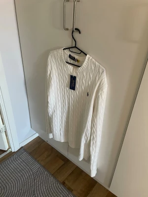 Vit kabelstickad tröja Polo Ralph Lauren - Kabelstickad vit tröja från Polo Ralph Lauren med rund halsringning och diskret broderad logga på bröstet. Tröjan har långa ärmar och ribbade muddar vid ärmslut och nederkant. Klassisk och stilren design i mjukt material.