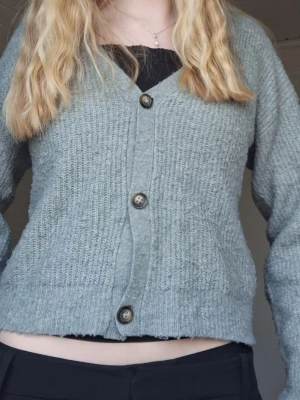 Grön stickad kofta med knappar - Mysig grön stickad kofta med v-ringning och fyra stora knappar framtill. Koftan har en rak passform, långa ärmar och ribbade muddar vid ärmslut och nederkant. Perfekt att slänga över en topp för en avslappnad look. Den är från Lager 157 och är i stl 160, passar även xs. Det är ganska nopprig men annars fin!