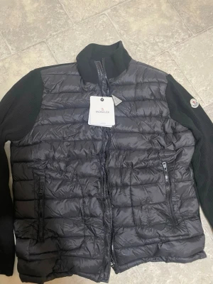 Svart dunjacka från Moncler - Snygg svart dunjacka från Moncler med klassisk Moncler-logga på ärmen och ett unikt serietidningsfoder på insidan. Jackan har en quiltad design och är tillverkad i glansigt material med dunfyllning som håller dig varm under kalla dagar. Stl 6 