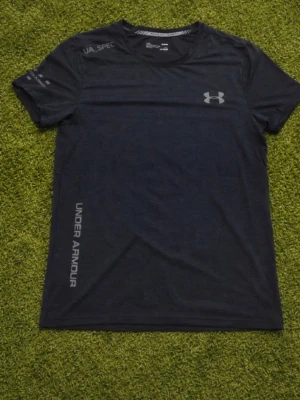 Svart träningströja från Under Armour - Snygg svart träningströja från Under Armour med reflekterande logga på bröstet och tryck på ärm och nederkant. Tillverkad i ett lätt och snabbtorkande funktionsmaterial som passar perfekt till gymmet eller löpning.