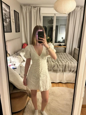 Vit kort klänning med spets och puffärm - Söt vit kort klänning med v-ringning, puffärmar och dekorativa knappar framtill. Klänningen har spetsdetaljer och är figursydd med lätt utsvängd kjol. Perfekt för student 