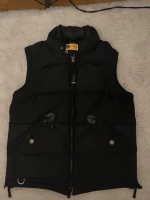Svart dunväst från Parajumpers - Snygg svart dunväst från Parajumpers Masterpiece Series med hög krage, dragkedja framtill och två snedställda fickor med dragkedja och tryckknapp. Västen har en gul detalj vid halsen och är perfekt för kalla dagar.