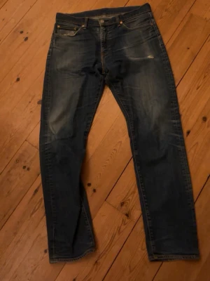 Mörkblå jeans från Levi's  - Säljer ett par klassiska mörkblå jeans från Levi's med raka ben och fem fickor. Jeansen har subtila slitningar framtill och en brun läderpatch bak i midjan. Perfekta till en avslappnad stil och passar till det mesta.