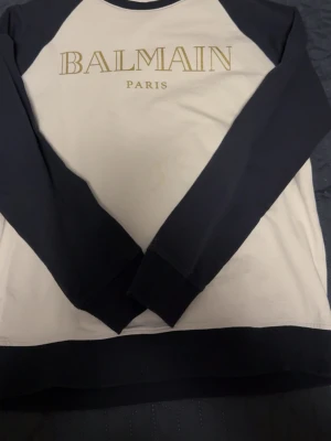 Svartvit sweatshirt från Balmain Paris - Snygg svartvit sweatshirt från Balmain Paris med guldfärgad logga framtill. Tröjan har svarta ärmar och nederkant, medan kroppen är vit. Tillverkad i mjukt bomullsmaterial och har en klassisk rund halsringning. Perfekt för dig som gillar exklusiva streetwear-plagg.