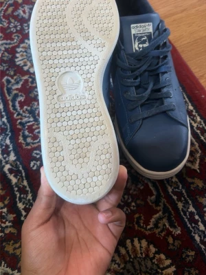 Adidas Stan Smith mörkblå sneakers - Snygga mörkblå Adidas Stan Smith sneakers med klassisk vit sula och diskret Adidas-logga på sidan och plös. Skorna har låga skaft och är tillverkade i slätt läder med matchande mörkblå snörning. Perfekta för en clean och stilren look.