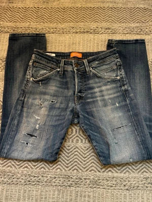 Blå slitna jeans från Jack & Jones - Säljer ett par blå jeans från Jack & Jones med slitningar och snygg tvätt framtill. Klassisk femficksmodell med raka ben och ljus tvätt på låren. Jeansen har en snygg vintage-look. De är i modell slim/glenn