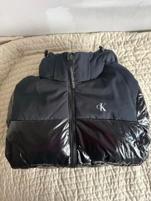 Svart pufferjacka från Calvin Klein - Snygg svart pufferjacka från Calvin Klein med blank finish på nedre delen och matt tyg upptill. Jackan har huva, dragkedja framtill och CK-logga på bröstet. Perfekt för kalla dagar och ger en modern streetwear-look.