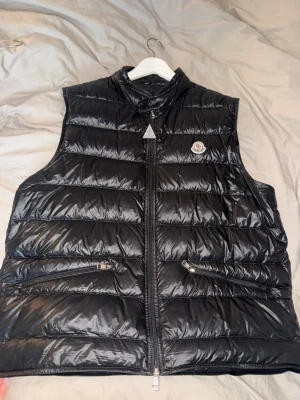 Svart dunväst från Moncler - Snygg svart dunväst från Moncler med glansig finish och klassisk logotyp på bröstet. Västen har hög krage, två dragkedjeförsedda fickor och quiltad design. Perfekt för lager-på-lager och streetwear-vibe.