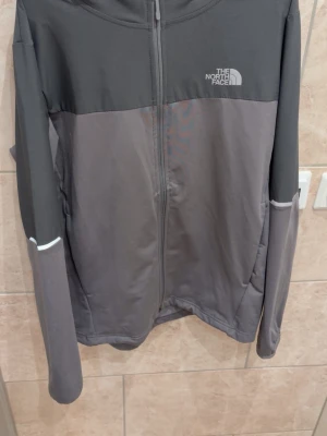Grå vindjacka från The North Face - Snygg tvåtonad grå vindjacka från The North Face med hel dragkedja framtill och diskret logga på bröstet. Jackan har långa ärmar och är tillverkad i ett lätt och smidigt material, perfekt för lager-på-lager och aktiva dagar.