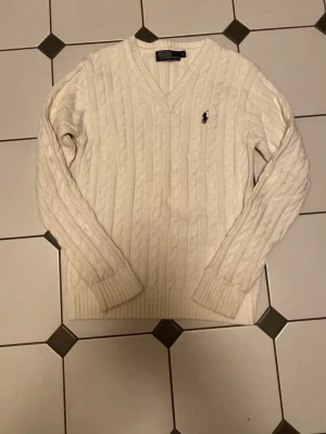 Vit kabelstickad tröja Polo Ralph Lauren - Vit kabelstickad tröja från Polo Ralph Lauren med V-ringning och broderad logga på bröstet. Tröjan har långa ärmar och ribbade muddar vid ärmslut och nederkant. Perfekt för dig som gillar klassisk och stilren design.