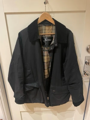 Barbour Bedale  - Svart Barbour Bedale med klassiskt rutigt innerfoder och krage i manchester. Jackan har stora fickor framtill, stängs med både dragkedja och knappar och har en tidlös, rak passform. Skicket är bra, men det finns en liten defekt på ryggen (syns knappt), saknas en knapp framtill och en inoljning kan behövas för den petiga. Storlek: M. Allt gott! /David