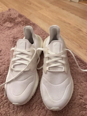 Vita Adidas Ultraboost sneakers - Snygga helvita Adidas Ultraboost sneakers med stickad ovandel i mesh och klassiska tre ränder på sidan. Skorna har en chunky sula för extra komfort och är tillverkade med återvunnen plast. Perfekta för dig som gillar stilrena och sportiga sneakers.