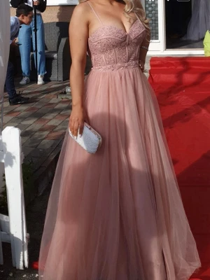 Lång rosa klänning med spetsdetaljer - Elegant långklänning i ljusrosa med spetsdetaljer på överdelen och tunna axelband. Kjolen är i flera lager av tunt, svepande tyg som ger ett romantiskt intryck. Perfekt för bal eller student. Klänningen har v-ringad front och markerad midja.