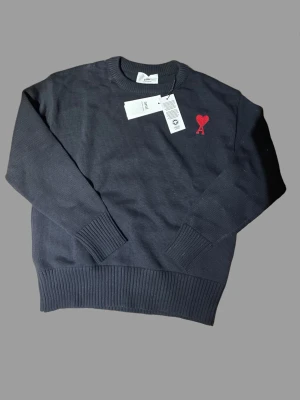 Svart stickad tröja från Ami Paris - Snygg svart stickad tröja från Ami Paris med röd broderad logga i form av ett hjärta och bokstaven A på bröstet. Tröjan har rund hals, ribbade muddar och nederkant. Perfekt för dig som gillar stilrena plagg med en twist.