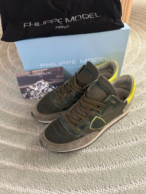 Philippe Model sneakers i grönt och gult - Snygga sneakers från Philippe Model i grönt och grått med detaljer i gult. Ovandelen är i en mix av mocka, läder och mesh för en cool och sportig look. Skorna har bruna snören och en låg silhuett. Perfekta för dig som gillar streetstyle och unika detaljer.