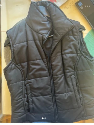 Svart quiltad dunväst med hög krage - Stilren svart dunväst med quiltat mönster och hög krage. Västen har dragkedja framtill och två fickor på sidorna. Perfekt för lager-på-lager och ger en sportig look. Materialet är mjukt och vadderat för extra värme.