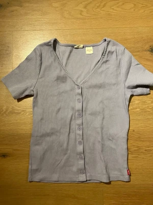 Ljuslila ribbad topp från Levi's - Supersöt ljuslila topp från Levi's med ribbad struktur och v-ringning. Toppen har korta ärmar och knappar hela vägen framtill. Perfekt att styla med jeans eller kjol för en chill look.