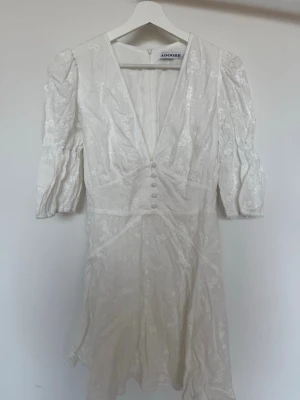 Sicily klänning från Adoore - Supersöt vit kortklänning från Adoore med v-ringning, puffärmar och knappar framtill. Aningen mindre i storlek. Mer som en 38a i midjan :)