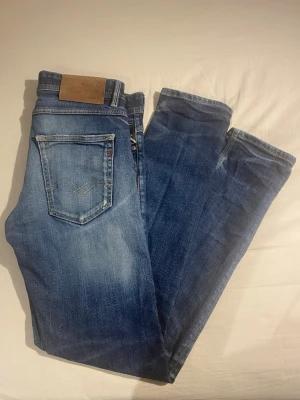 Blå jeans från Replay - Snygga blå jeans från Replay med klassisk femficksmodell och ljusa slitningar. Jeansen har raka ben och detaljerade sömmar på bakfickorna samt en läderpatch med logga i midjan. Perfekta till en avslappnad streetstil. W28-L32