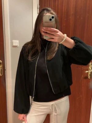 Svart bomberjacka med dragkedja - Stilren svart bomberjacka med rund halsringning och silverfärgad dragkedja framtill. Jackan har ballongärmar och resår i midjan för en snygg passform. Är i storlek M men är liten i storleken, så sitter ungefär som en S😊