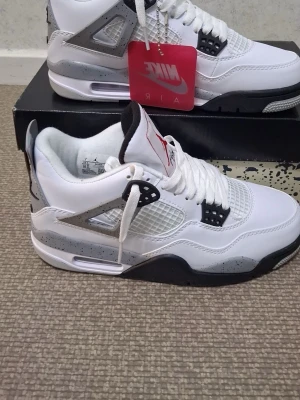 Nike Air Jordan 4 White Cement - Säljer ett par Nike Air Jordan 4 White Cement sneakers. Skorna är vita med grå och svarta detaljer samt röda inslag på sulan. Klassisk meshpanel på sidan, plastdetaljer och synlig Air-enhet i sulan. Ikonisk Jumpman-logga på tungan och hälen. Perfekta för dig som gillar streetwear.