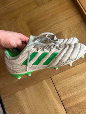 Vita och gröna Adidas fotbollsskor - Säljer ett par vita fotbollsskor från Adidas med gröna detaljer och klassiska tre ränder på sidan. Skorna har snörning, grönt foder och dobbar för spel på gräs. Ovandelen är i syntetmaterial med mönstrad struktur för bättre bollkontroll.