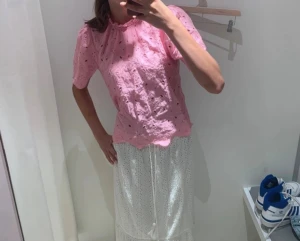 Rosa broderad blus med puffärm - Söt rosa blus med broderade hål och puffiga korta ärmar. Blusen har liten volang vid halsen och en knapp i nacken. Perfekt för en trendig och feminin look. Materialet känns lätt och luftigt.