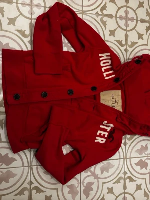 Röd hoodie från Hollister med knappar - Snygg röd hoodie från Hollister med stora svarta knappar framtill och broderad vit logga på bröstet. Tröjan har huva med dragsko och två fickor framtill. Hör av er vid frågor🤝😁