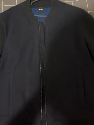 Mörkblå bomberjacka från BOSS - Snygg mörkblå bomberjacka från BOSS med minimalistisk design. Original pris 4000 kr säljer den för 1300 kr för den är liten  Jackan har ribbade muddar, diskreta sidofickor och dragkedja framtill. Tillverkad i ett mjukt tyg som ger en clean och stilren look. Perfekt för dig som vill ha en klassisk och trendig jacka. 