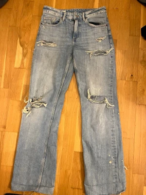 Ljusblå slitna jeans från Zara - Säljer ett par ljusblå jeans från Zara med raka ben och flera slitningar och hål för en trendig, avslappnad look. Jeansen har klassisk femficksmodell och är tillverkade i bomull. Perfekta för dig som gillar streetstyle och en edgy vibe.