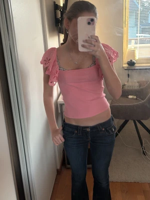 Rosa croppad topp med volangärmar - Söt rosa croppad topp med stora volangärmar och blommiga detaljer. Toppen har en fyrkantig halsringning och är ärmlös med dekorativa axelvolanger. Perfekt för dig som gillar färg och unika detaljer.